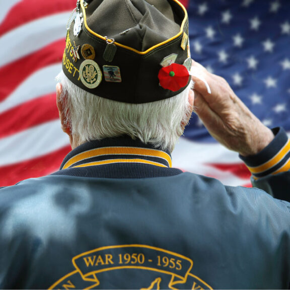 photo of the back of a Veterans wearing veterans jacket and hat saluting the American flag