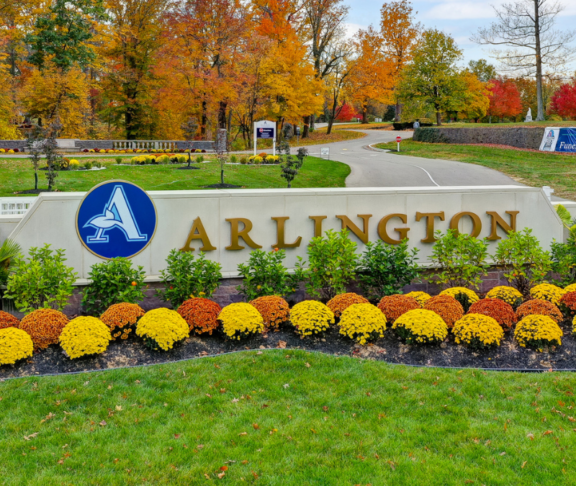 Arlington Front Gate with a blue logo featuring the letter 'A', surrounded by vibrant yellow and orange flowers. A curving road, autumn trees, and other signage are visible in the background.